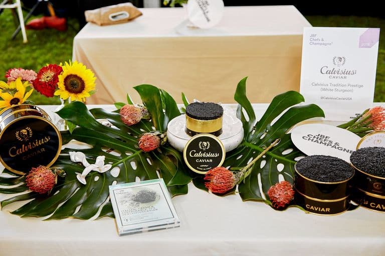 Elegant caviar tasting display featuring Calvisius Caviar tins arranged on tropical leaves with decorative flowers, showcasing luxury caviar varieties at a gourmet food and champagne event.
