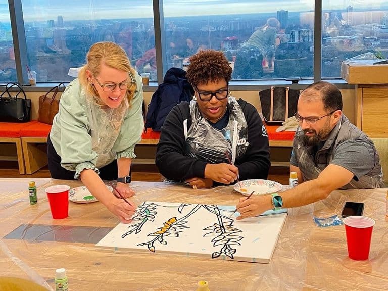 Three coworkers painting a colorful mural together at a bright office table with city views behind them.
