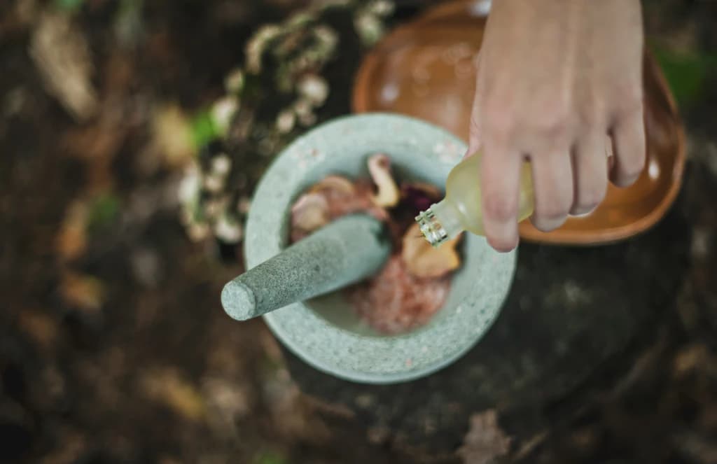 A person pouring herbal liquid into a bowl while using a mortar and pestle, set outdoors on a wooden surface.