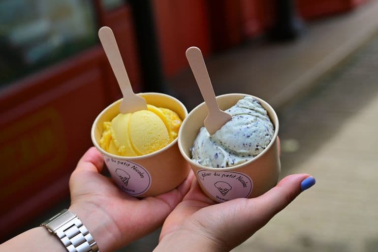 Two hands holding paper cups of ice cream—one with yellow scoops, the other with cookies and cream—each with a wooden spoon.