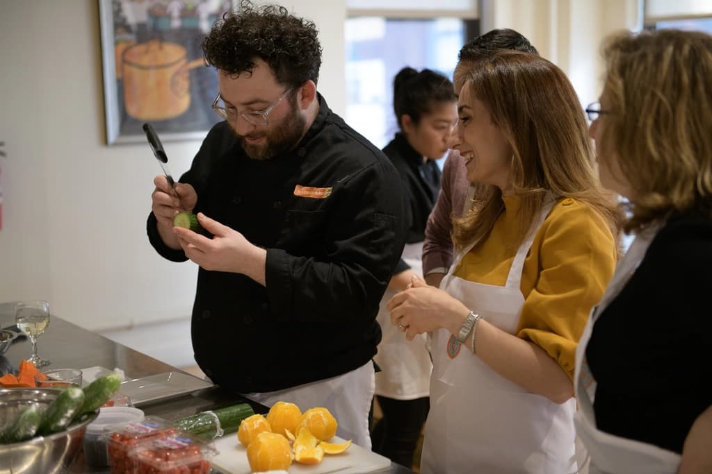 A chef demonstrating a technique in a cooking class, surrounded by participants watching attentively, with fresh ingredients on the table.