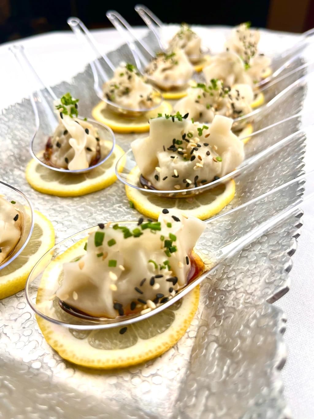 A close-up of elegantly plated dumplings garnished with sesame seeds and herbs, served on lemon slices with soy sauce on clear spoons.