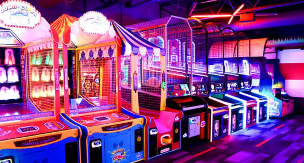 A vibrant arcade with neon lights showcasing various colorful games, including basketball hoops and carnival-style games, creating a lively atmosphere.