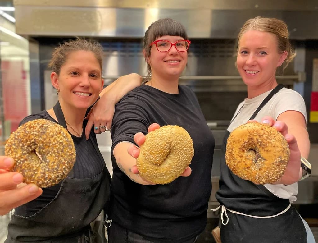 Three smiling bakers holding freshly baked bagels in front of them, each showcasing a different type of bagel in a kitchen setting.