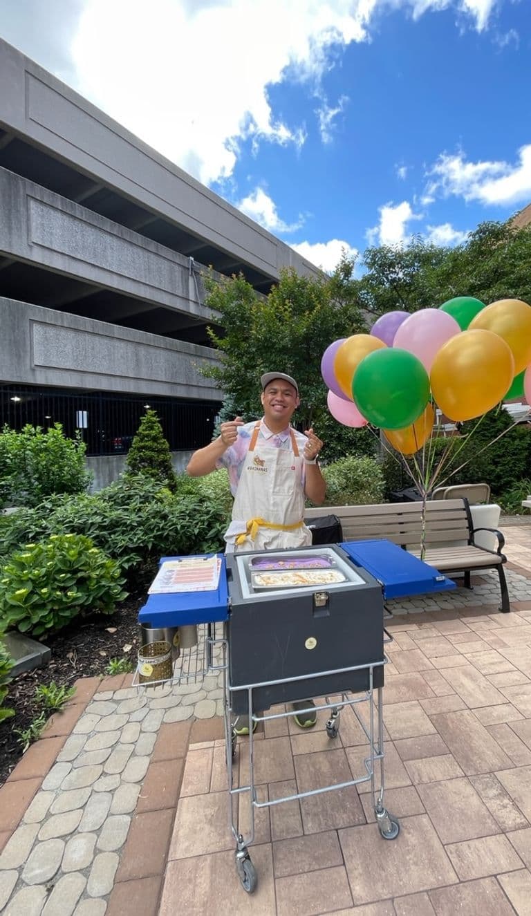 Smiling vendor standing behind a rolling banana pudding cart with colorful balloons in an outdoor plaza surrounded by greenery and buildings.