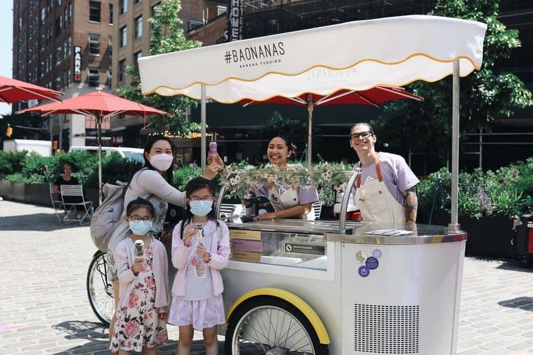 Outdoor banana pudding cart with two smiling vendors serving a masked woman and two children holding ice cream treats, set in a sunny urban plaza.