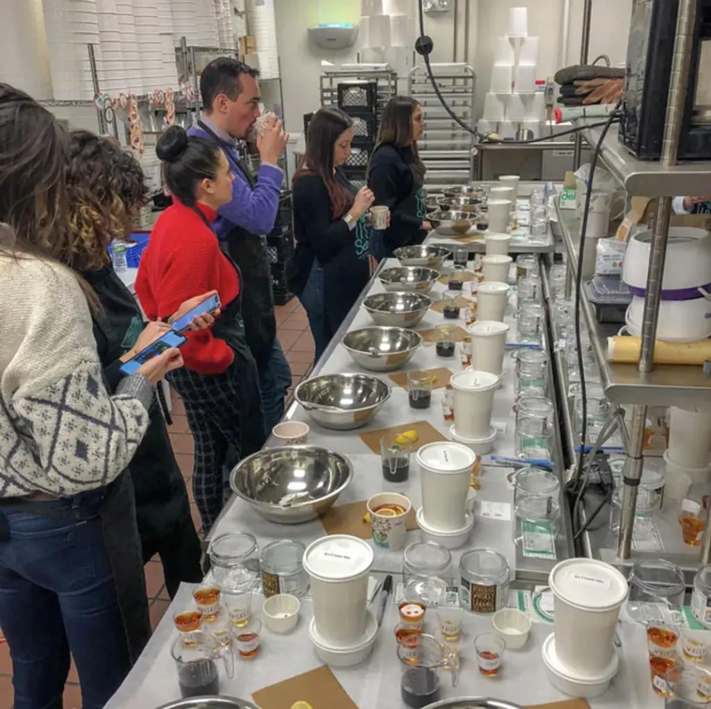 A group of people smiling and raising their glasses for a toast at a boozy ice cream-making event, gathered around a table of ingredients.