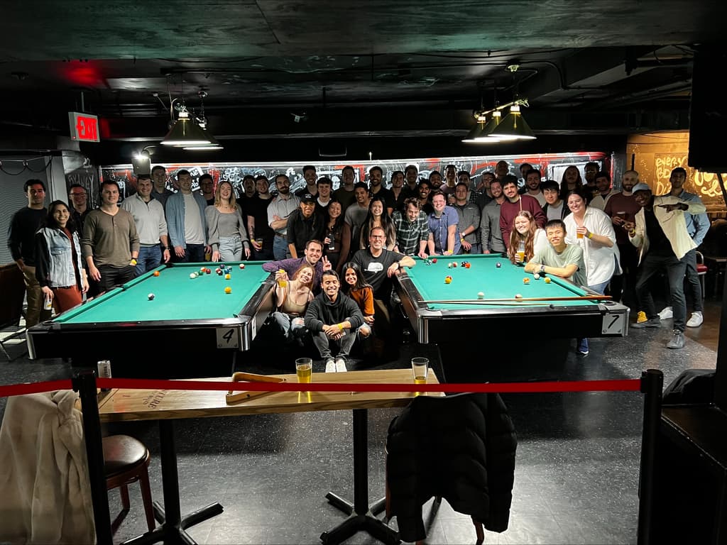 A large group of people gathered around pool tables in a dimly lit room, smiling and posing for a photo.