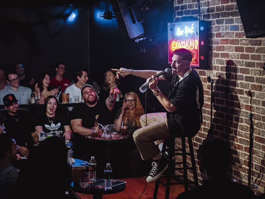 a comic on a stage with a microphone in hand in front of a crowd of people laughing