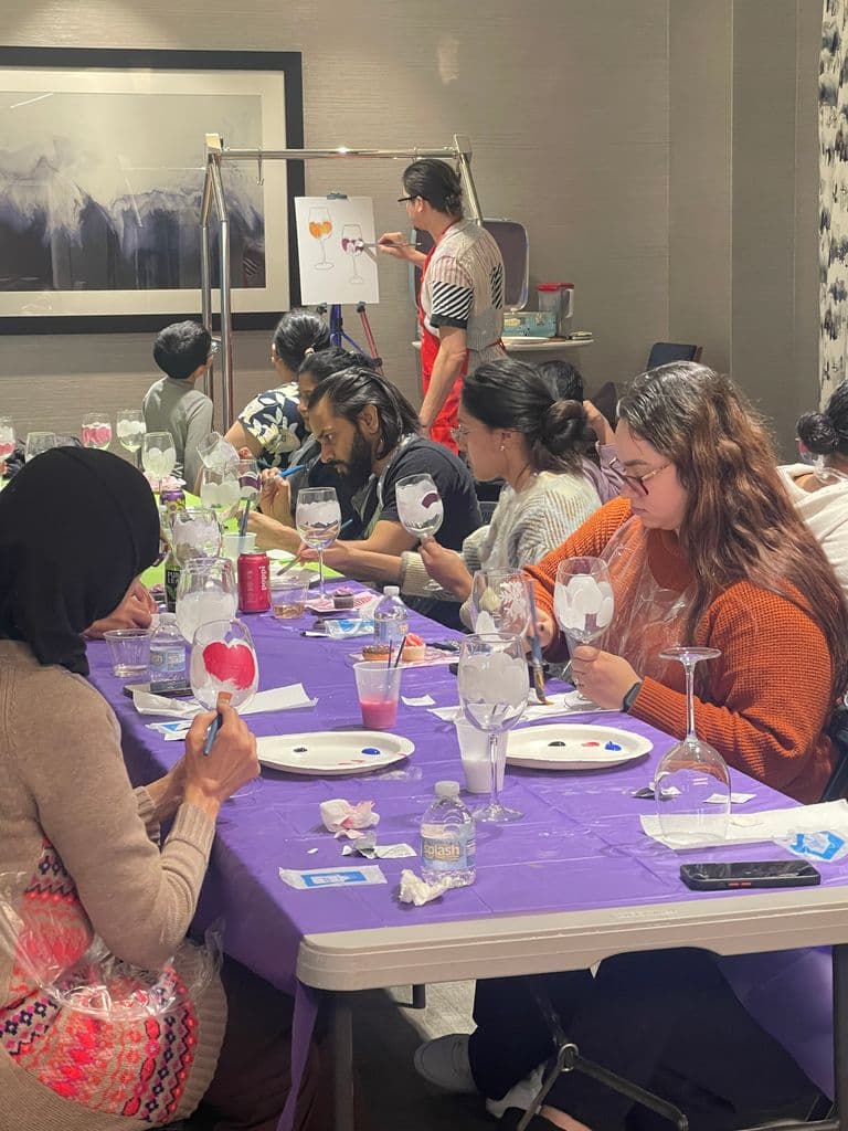 A group painting wine glasses at a guided art event, following an instructor's example. The setting has a creative and social atmosphere.