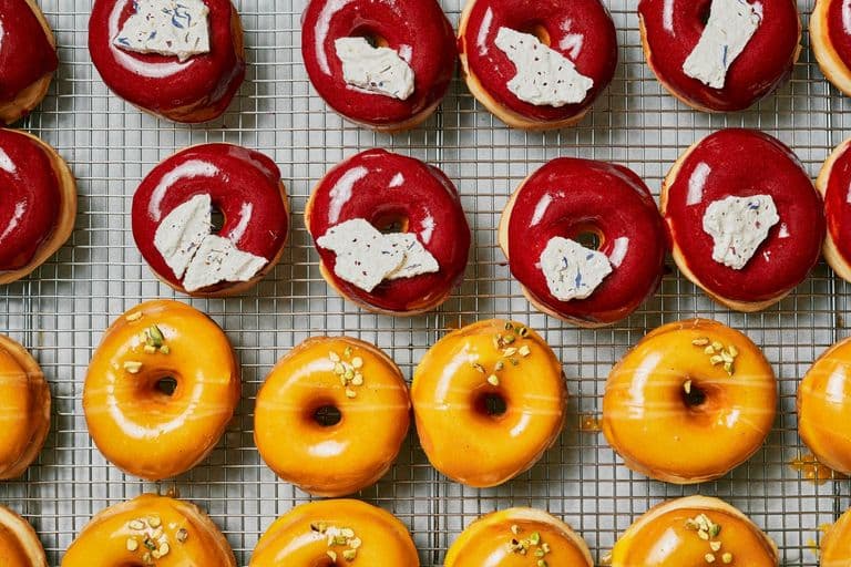 Rows of glossy red and orange glazed doughnuts on a cooling rack, topped with cream, pistachios, and decorative garnishes