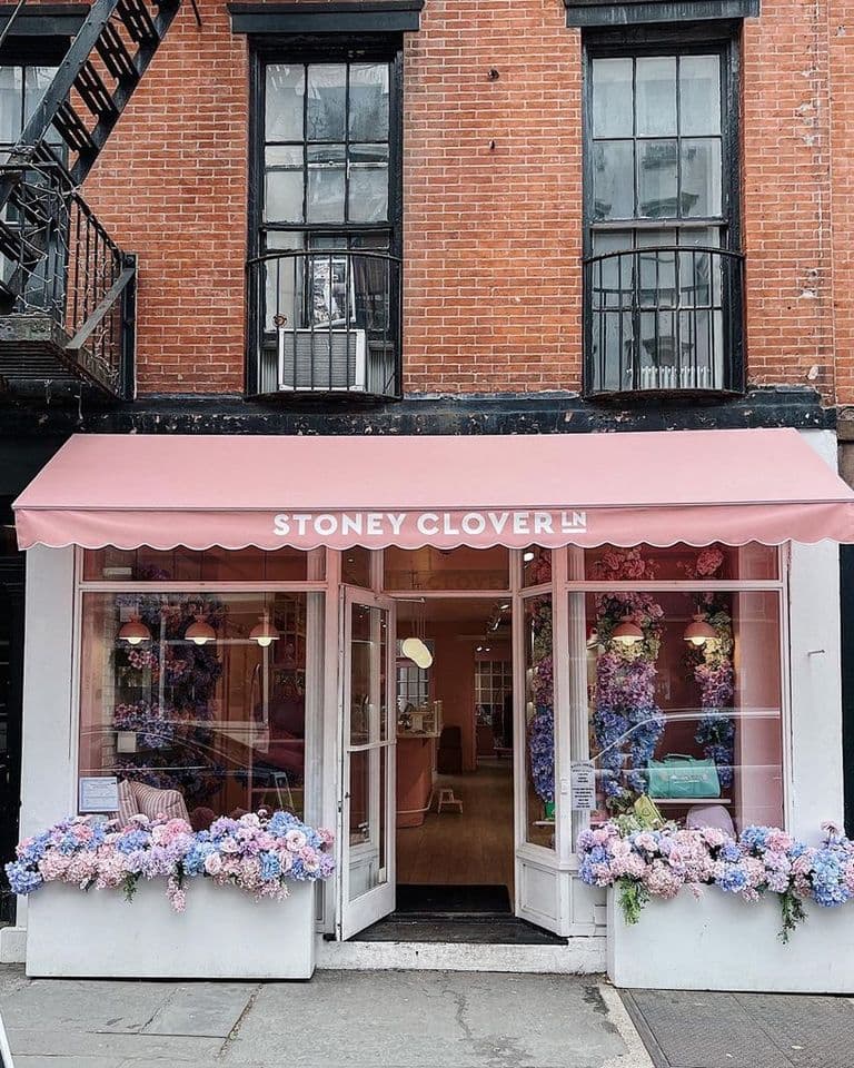 Exterior of the Stoney Clover Lane storefront featuring an open glass door and large display windows. The entrance is decorated with pastel pink flowers, white chrysanthemums, and an assortment of neutral-toned pumpkins, creating a cozy, seasonal look. Inside, soft pink walls and colorful bags are visible, adding to the boutique’s whimsical charm. The storefront is set in a classic brick building with black trim and iron fire escapes above.