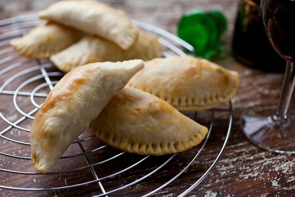 Freshly baked empanadas cooling on a wire rack, with a glass of red wine and a green bottle in the background, creating a rustic and cozy ambiance.