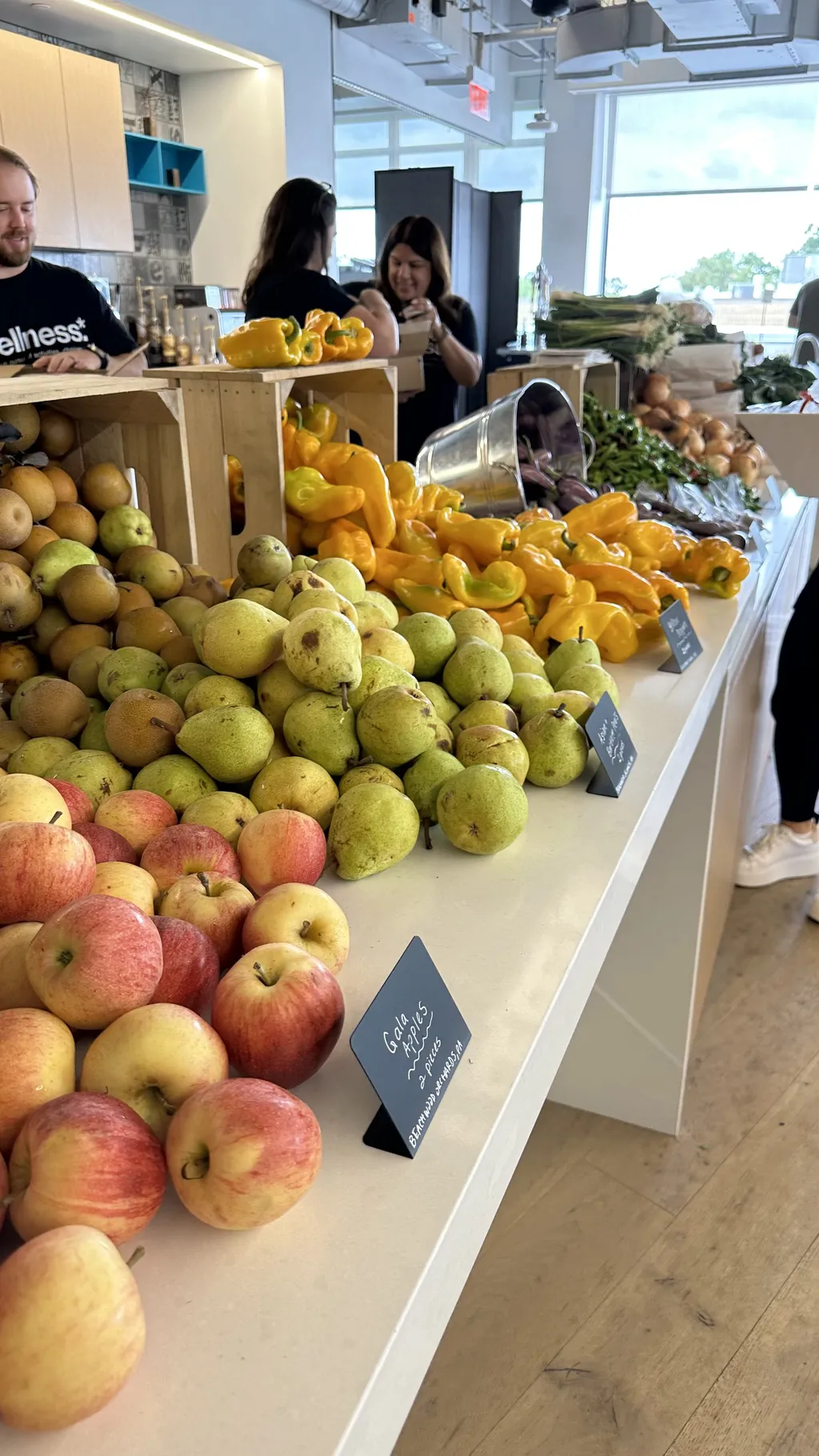 A vibrant array of fruits and vegetables, including apples, pears, and yellow bell peppers, neatly displayed on a table at an indoor farmer's market, with customers browsing in the background.