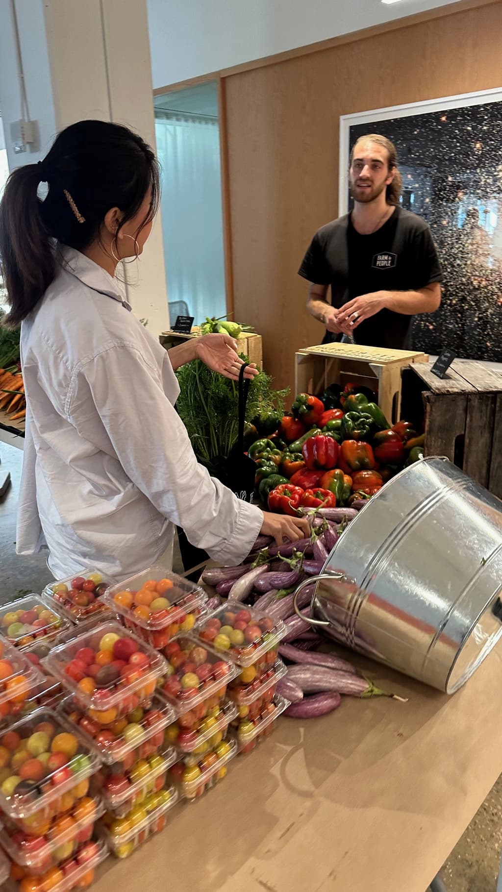 A customer at a farmer's market selecting fresh produce from a stand filled with colorful vegetables like peppers, eggplants, and cherry tomatoes, interacting with a vendor.