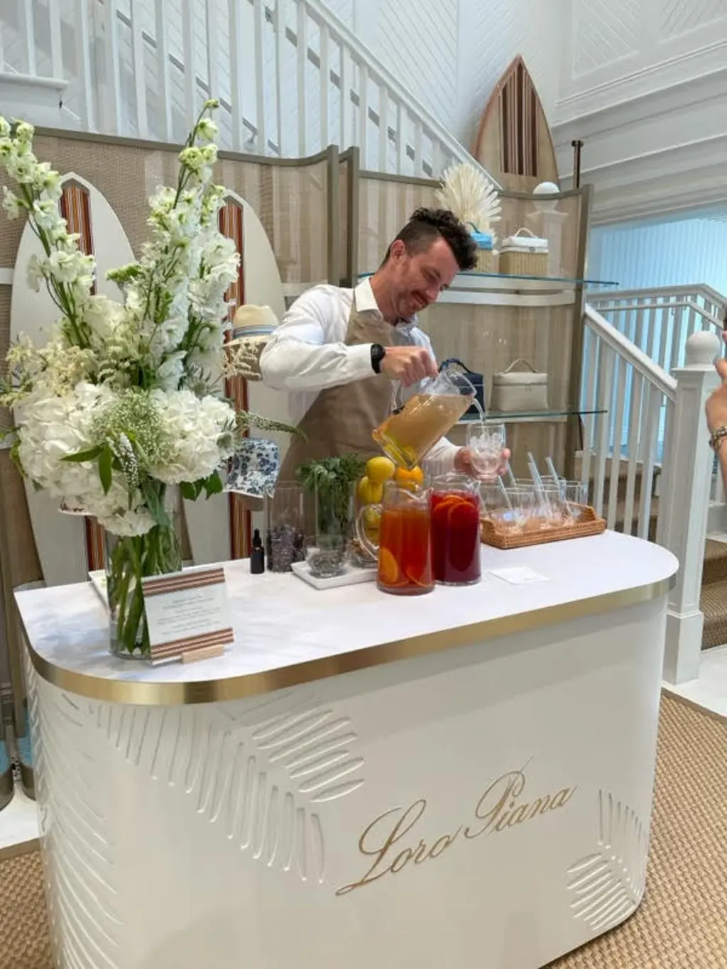 A man in an apron pours colorful drinks from a pitcher at a white bar counter decorated with fresh flowers inside a bright, elegant space.