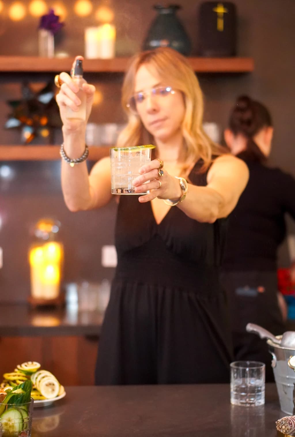 A woman wearing glasses sprays a cocktail with a mist while holding the glass in a stylish bar setting.