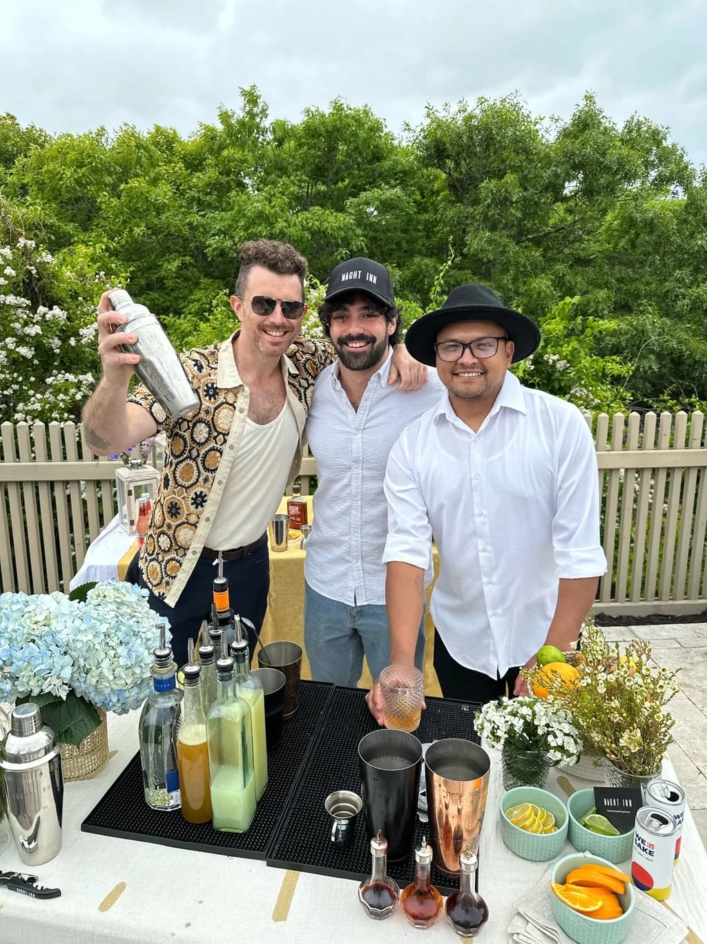Three men standing behind a cocktail station outdoors, smiling at the camera. The station is set with bottles, shakers, and garnishes under a cloudy sky.