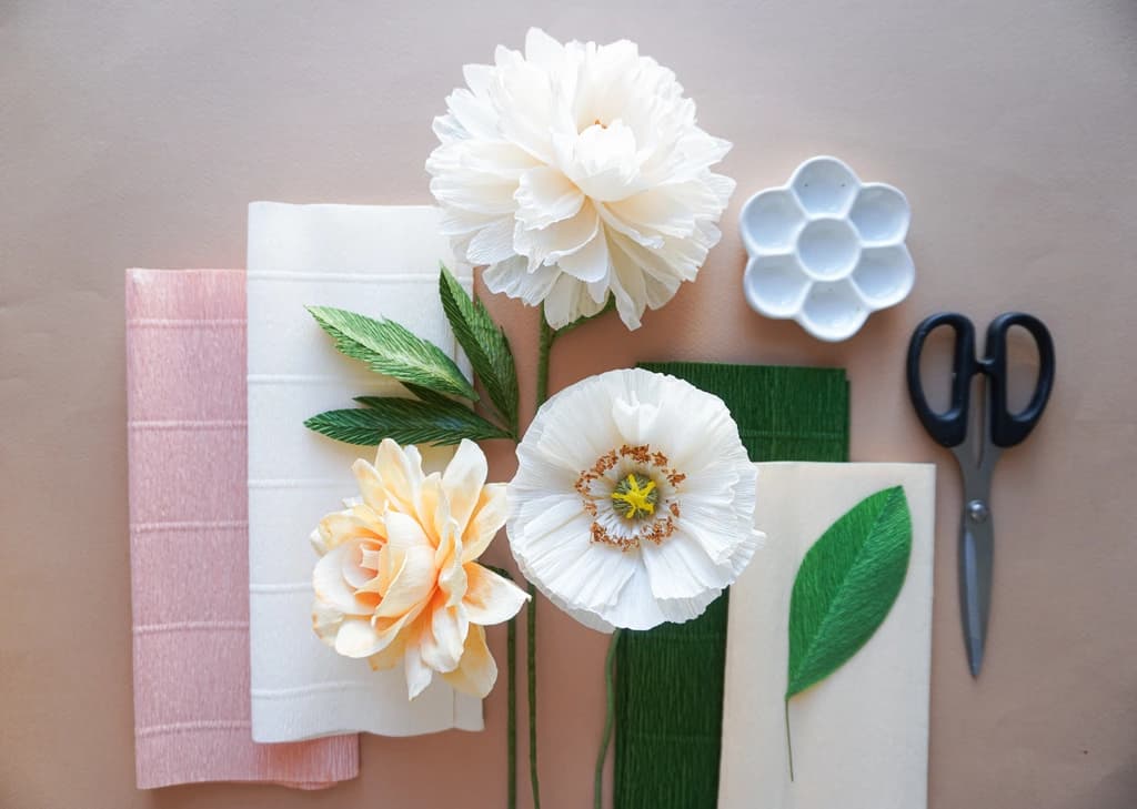 A flat lay of crafting supplies, including white and pastel-colored paper flowers, green leaves, scissors, and crepe paper sheets, ready for assembly on a neutral background.