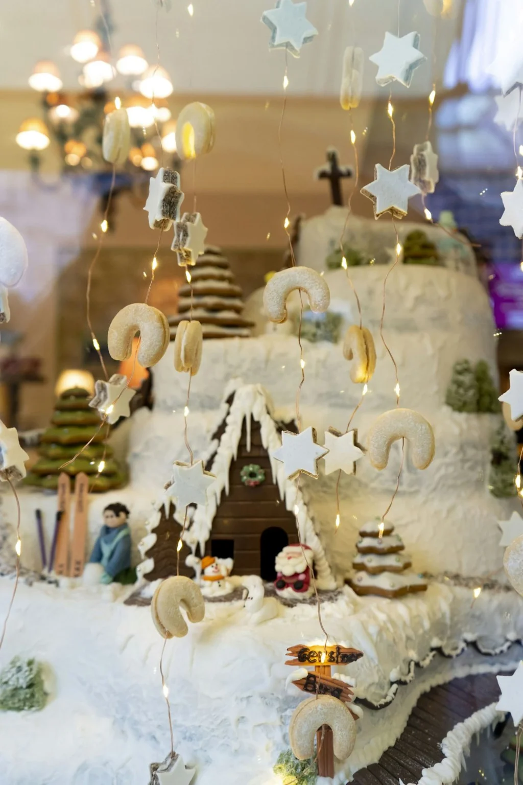 A gingerbread village display with icing-covered houses, holiday figurines, and star-shaped hanging cookies, illuminated by warm twinkle lights.