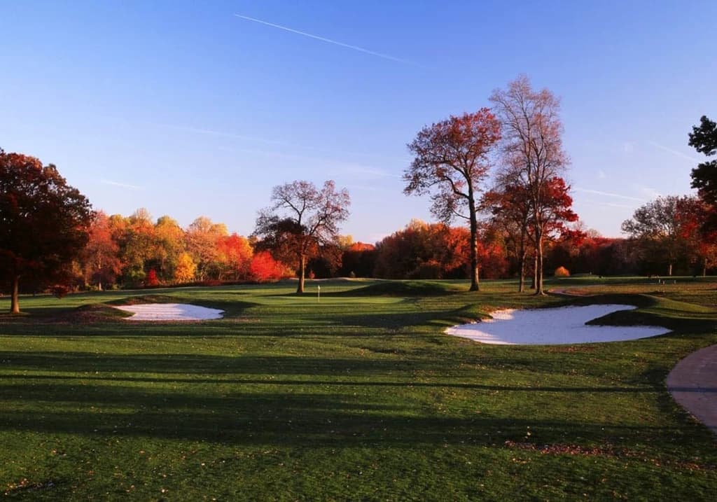 A scenic view of a golf course with sand bunkers, trees with autumn foliage, and a clear blue sky.