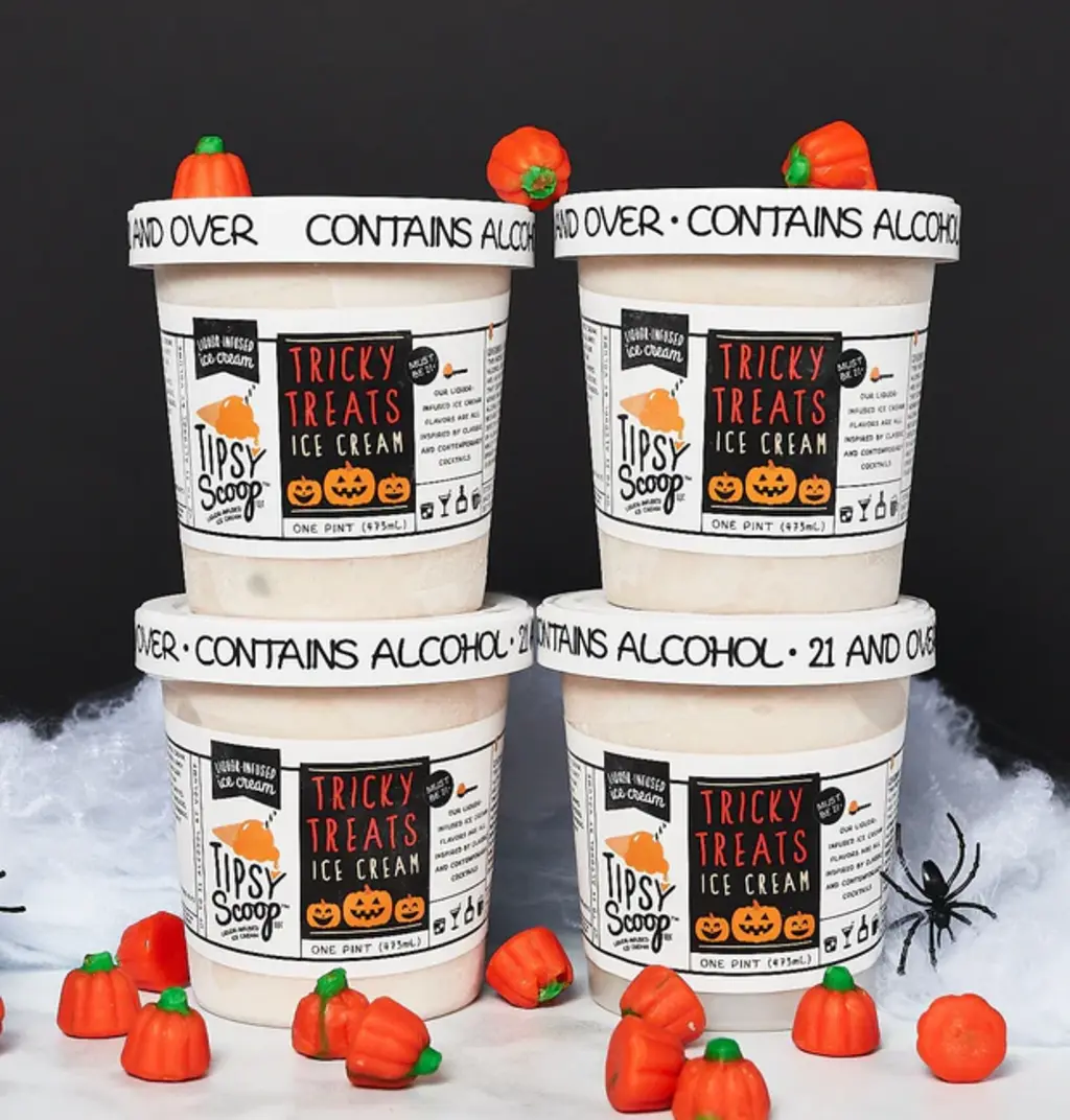 Four pints of Tipsy Scoop Tricky Treats ice cream, stacked with mini pumpkin candies, decorated for Halloween on a black background.