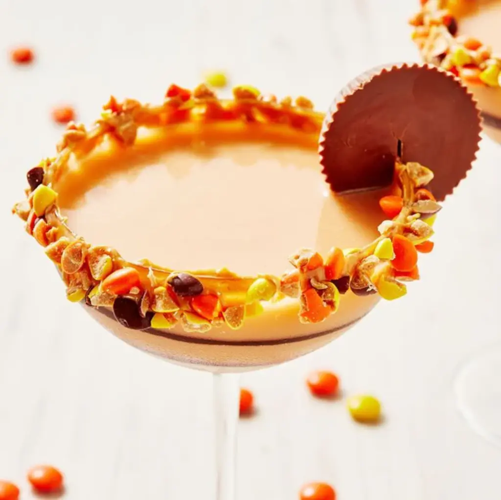 Four mini ice cream cups topped with Halloween candies and a Butterfinger bar, displayed in a wooden tray in front of a Tipsy Scoop window.