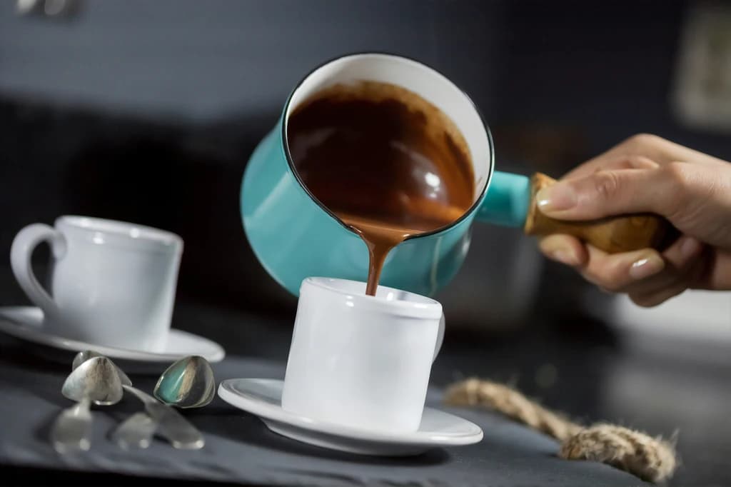 Pouring rich, thick hot chocolate from a turquoise pot into a small white cup.