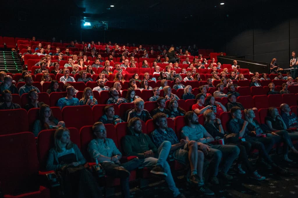A packed movie theater with an audience watching a film, seated in red chairs under dim lights.
