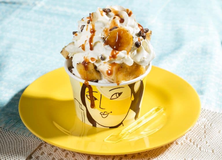 Ice cream sundae with whipped cream, caramel drizzle, and caramelized banana pieces in a branded cup on a yellow plate.