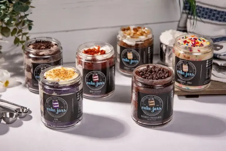 Assorted cake jars in various flavors, including ube, red velvet, and double chocolate, displayed on a white table with measuring spoons and decorative greenery.