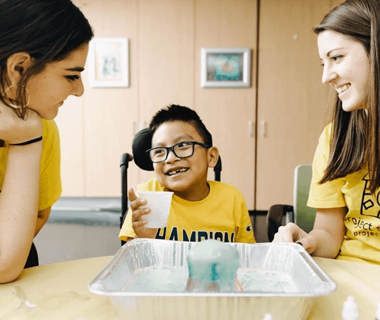 Child smiling with two adults during a hands-on science activity, wearing matching yellow shirts.