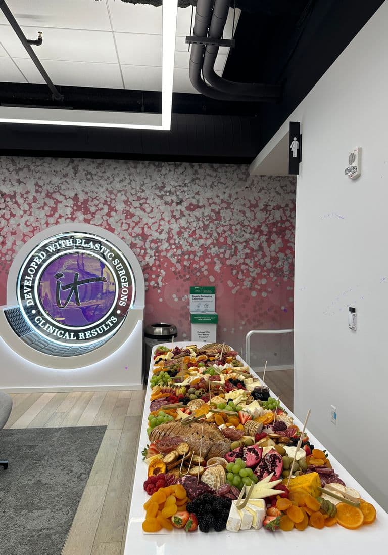 Office lobby with a long white table showcasing an abundant grazing spread of cheeses, meats, fruits, crackers, and bread, set against a pink and white decorative wall.