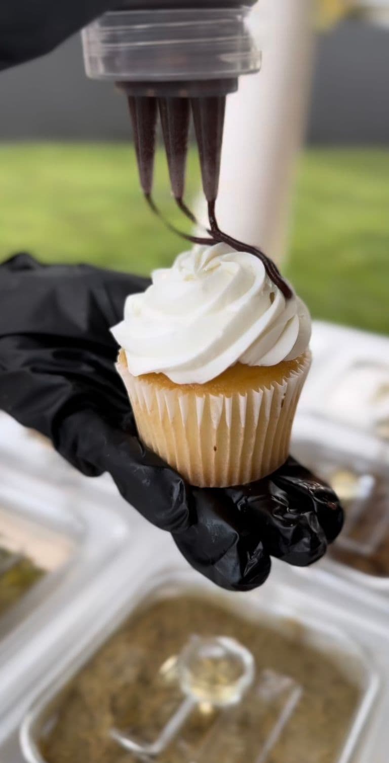 Vanilla cupcake with piped whipped frosting as chocolate drizzle is added by hand at a dessert topping station.
