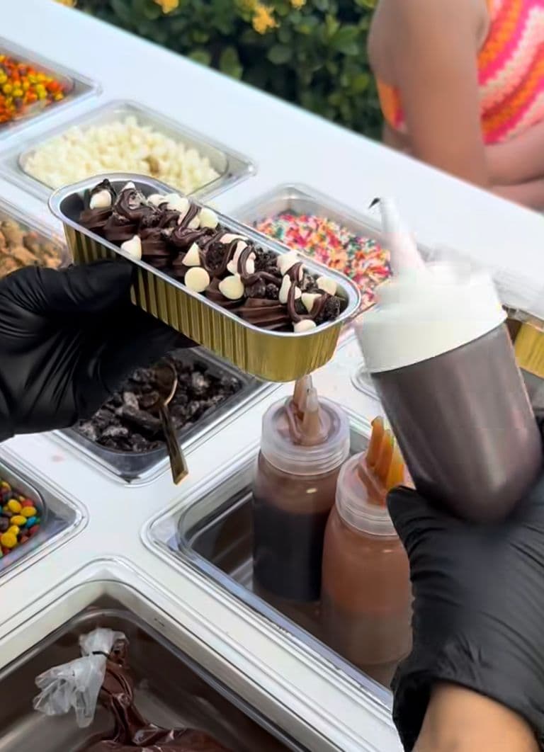 Chocolate loaf cake topped with chocolate frosting, mini marshmallows, cookie pieces, and drizzle at a dessert topping bar.