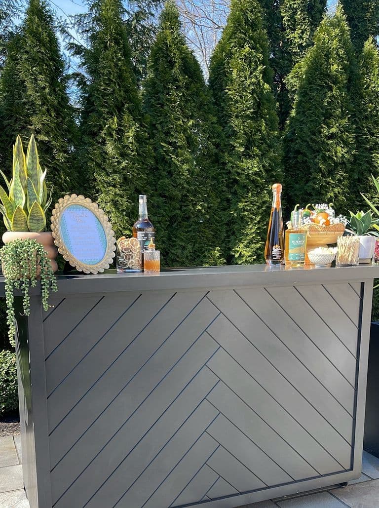 Outdoor bar setup with a sleek gray chevron-patterned bar, decorated with potted plants, bottles of liquor, garnishes, and a framed drink menu, set against a backdrop of tall green trees.