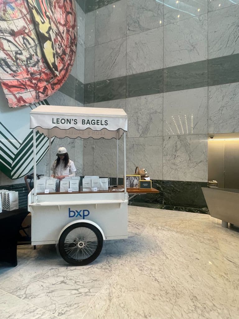 A white bagel cart labeled "Leon's Bagels" is set up in a modern lobby with marble walls, staffed by a woman in a cap and mask.