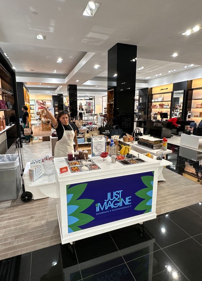 A person behind a mobile mini pancake bar inside a department store, surrounded by toppings and equipment, with a sign reading “Just Imagine Saturdays @ Bloomingdale’s."