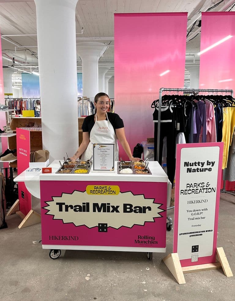 Person standing behind a colorful “Trail Mix Bar” station in a retail space, offering various mix-ins for custom trail mixes, with bright pink banners and clothing racks in the background.