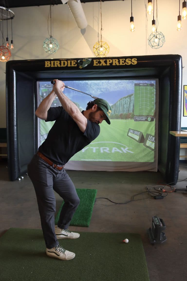 Person mid-swing with a golf club in front of an indoor golf simulator screen, set up in a modern event space with hanging lights.
