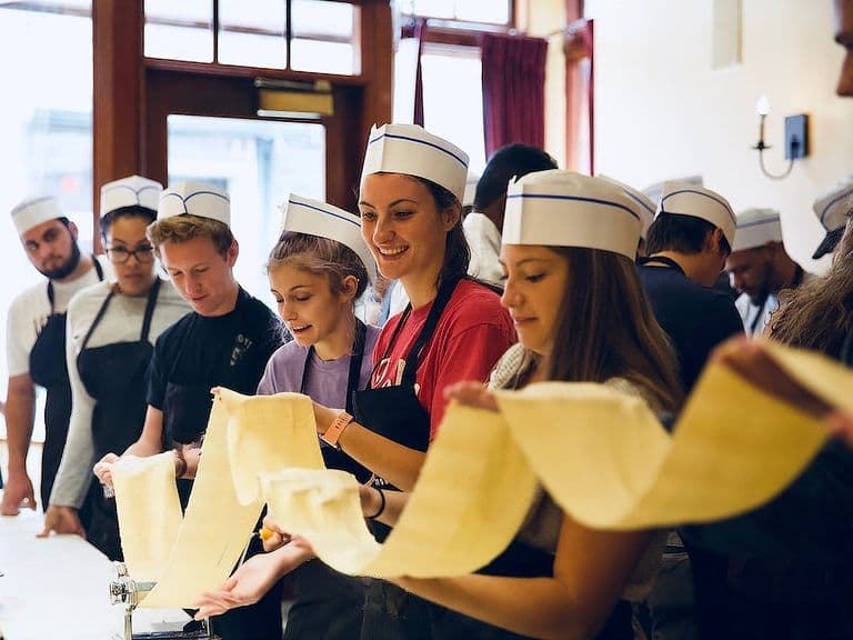 Group wearing chef hats smiles as they stretch fresh pasta sheets during a pasta-making workshop.