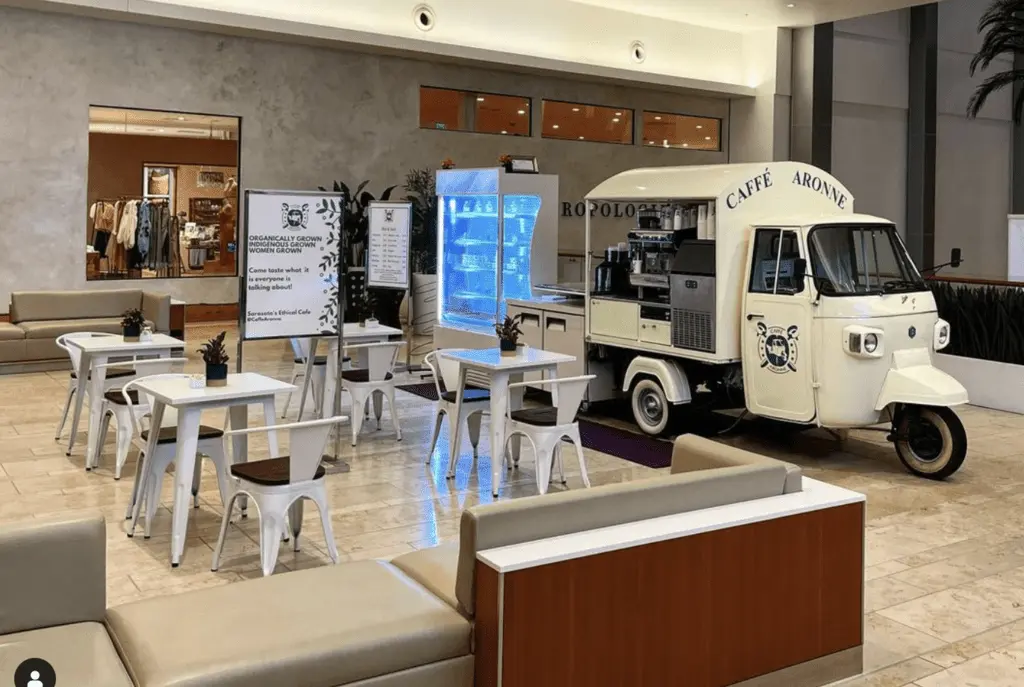 A coffee cart setup in a mall, featuring a white three-wheeled truck labeled Caffè Aronne, surrounded by tables and seating.
