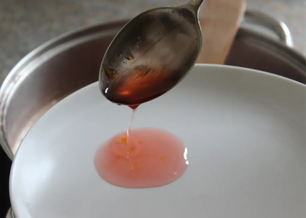 A spoon holding a small amount of red liquid being tested for consistency over a white plate. The spoon is slightly tilted, allowing the liquid to drip onto the plate.