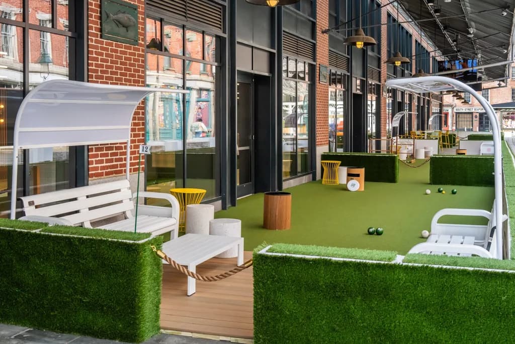 Outdoor seating area with artificial grass, white benches, and shaded canopies in front of a building.