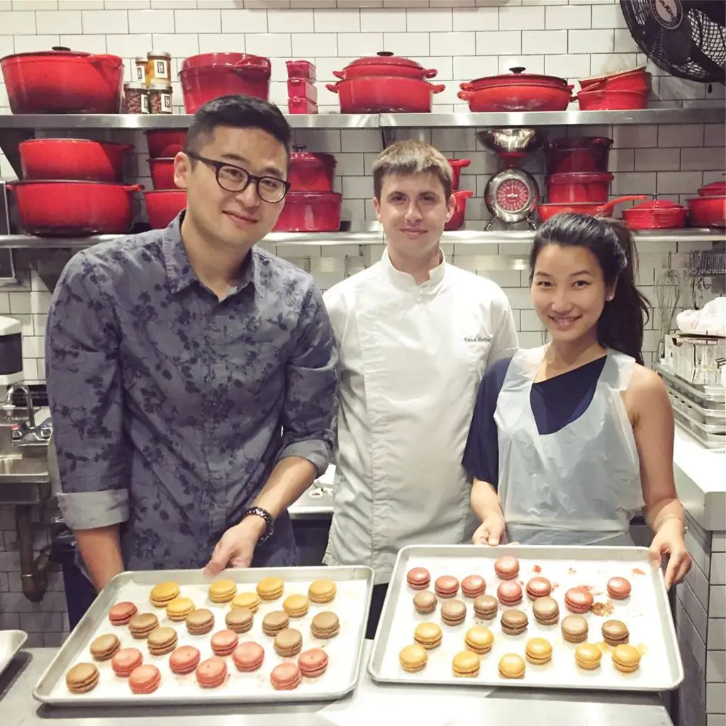 Three individuals in a kitchen posing with trays of colorful macarons, with red cookware displayed on shelves behind them.