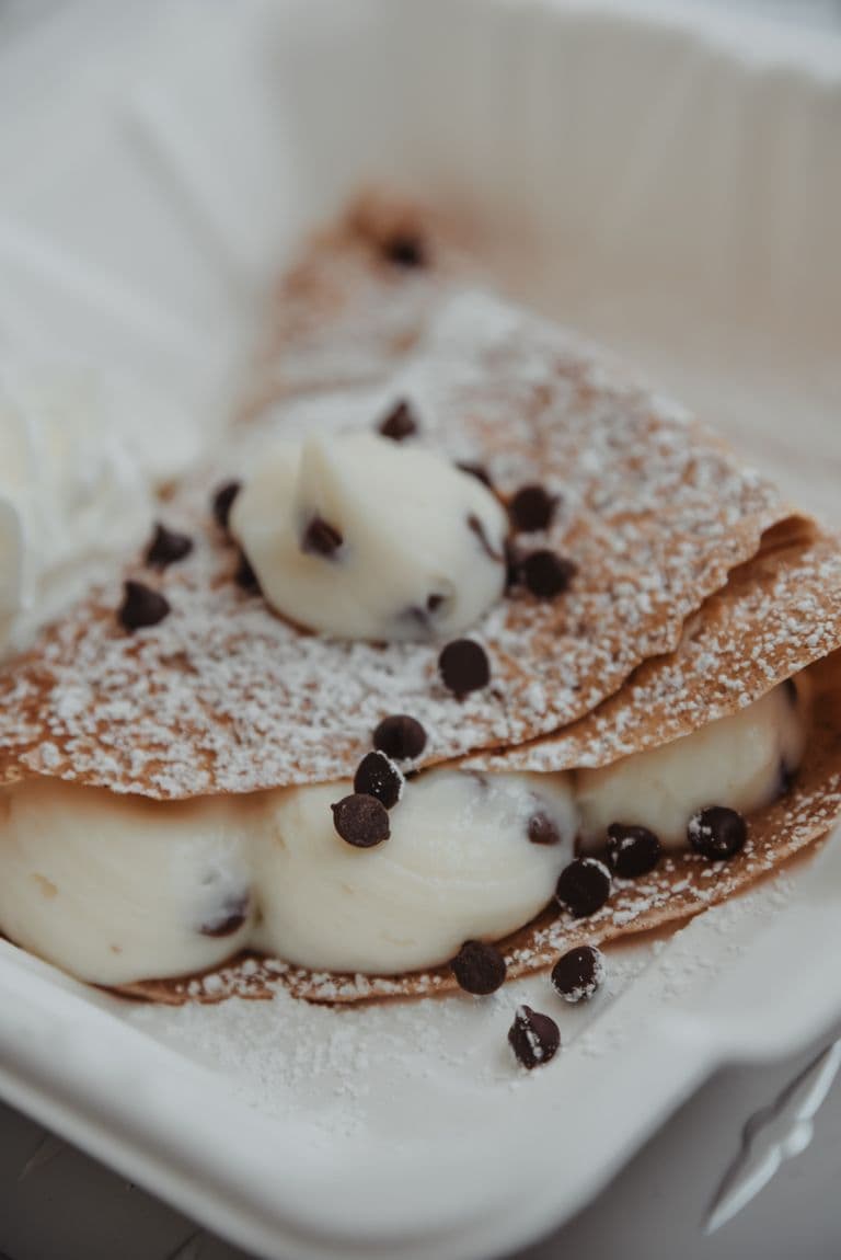 A golden crepe filled with cream and chocolate chips, dusted with powdered sugar, served in a takeout container.