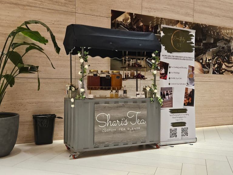 A stylish mobile tea bar setup with a black canopy and floral accents, displaying jars of tea and signage for “Shari’s Tea – Custom Tea Blends,” promoting tea tastings, tea mixology, and mobile café experiences.