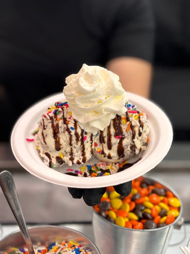 Two scoops of vanilla ice cream with chocolate drizzle, rainbow sprinkles, and whipped cream on top.