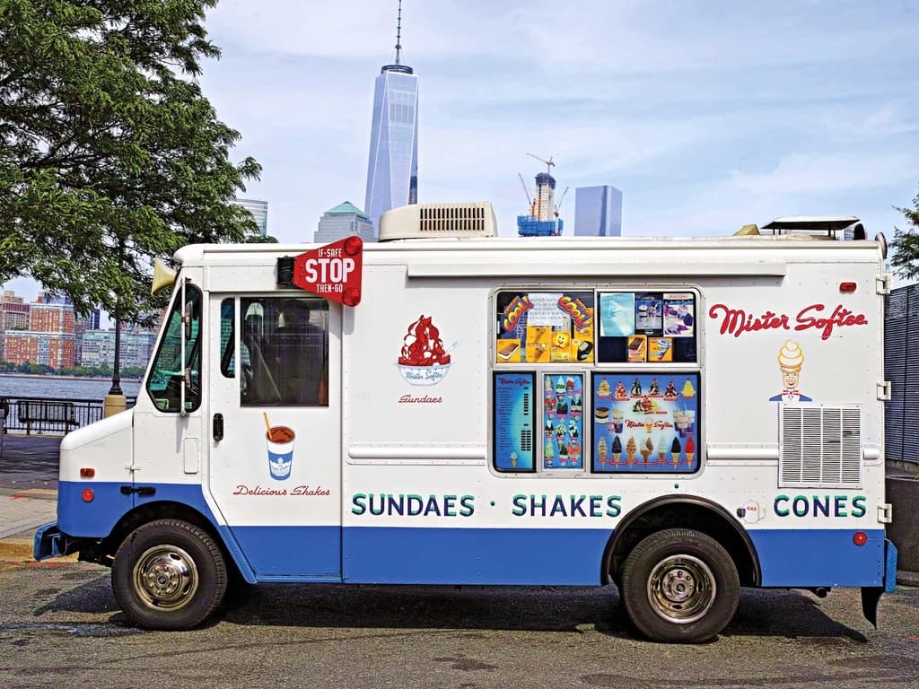 A classic Mister Softee ice cream truck parked on a city street, featuring colorful menu items on the side, including sundaes, shakes, and cones, with the city skyline in the background.