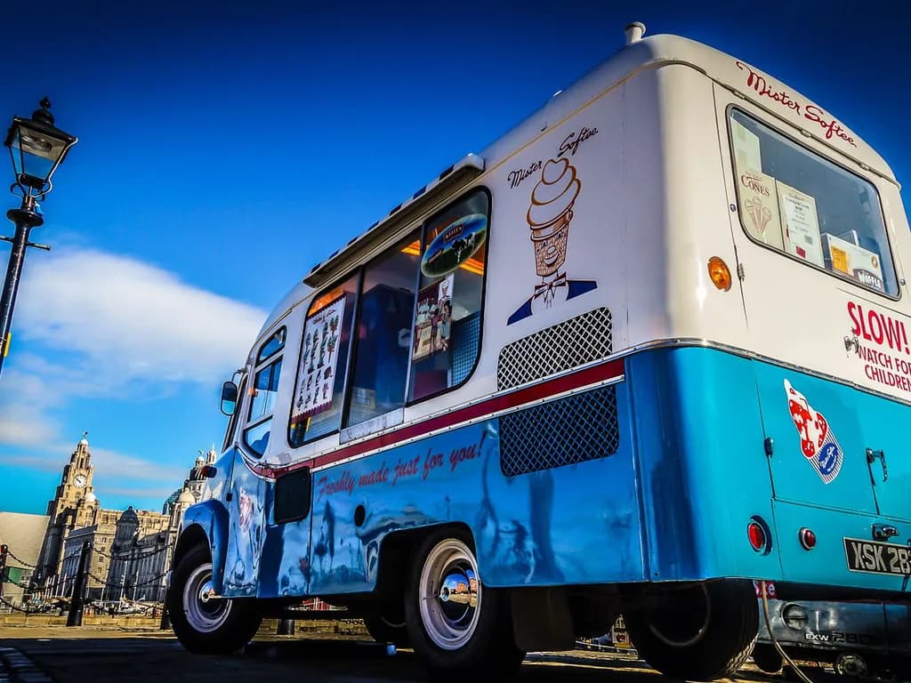 A classic Mister Softee ice cream truck with blue and white colors, featuring a large ice cream cone logo, a serving window, and menu items displayed on the side. The truck is parked with a clear blue sky and city buildings in the background.
